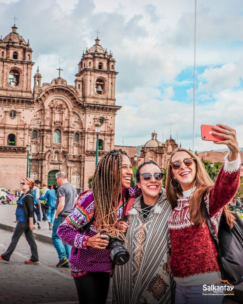 Three tourists in Cusco's main square smiling and taking a selfie with their friends.