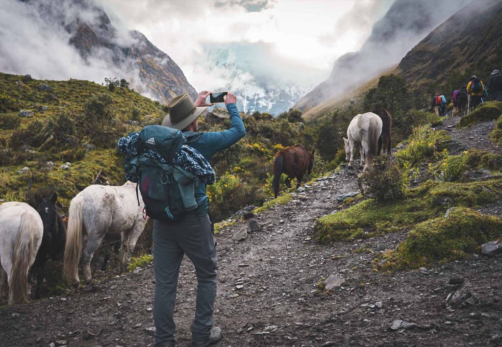 A tourist taking a photo with his cell phone on the Salkantay Trail, surrounded by vegetation, mist, and horses