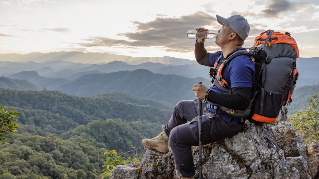 A hiker drinking water from a bottle while standing with two friends on a rocky mountain trail with snowy peaks.