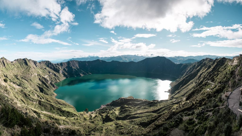 A panoramic view of the massive emerald green volcanic crater lake of Quilotoa under a cloudy sky.