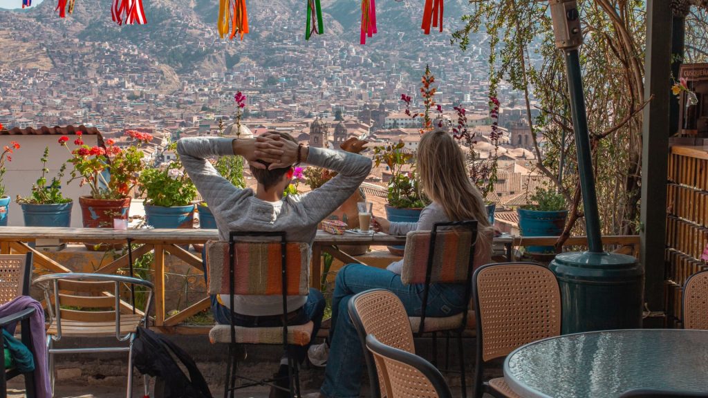 A scenic view of the vibrant Plaza de Armas in Cusco, Peru, framed by the wooden windows of a second-floor restaurant balcony terrace, showing the colonial architecture and mountains under a blue sky.