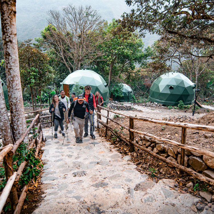 A diverse group of hikers with backpacks standing and talking on a wooden platform at the Super Jungle Domes campsite in Lucmabamba, surrounded by lush tropical vegetation and mountains